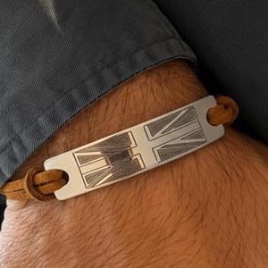Puede incluir: Una pulsera plateada con una correa de cuero marrón. La pulsera presenta el diseño de la bandera de la Union Jack. La correa de cuero se asegura a través de los agujeros de la pulsera. La pulsera mide aproximadamente 2,5 cm de ancho.