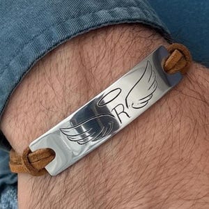 Può includere: Un braccialetto d'argento con una placca rettangolare con ali d'angelo e la lettera "R". Il braccialetto ha un cinturino in pelle marrone. Il disegno è inciso sulla placca metallica.