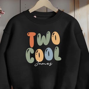 May include: A black long-sleeve sweatshirt featuring the text "TWO COOL" in large, rounded, multi-colored bubble letters in shades of orange, sage green, and light blue, with the name "James" written in a white cursive script font below.