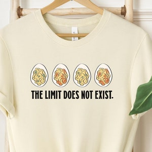 May include: Cream-colored t-shirt with the text "THE LIMIT DOES NOT EXIST." The shirt features a graphic of four eggs with different colored yolks. The shirt is hanging on a wooden hanger.