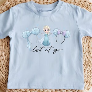K&ouml;nnte beinhalten: Hellblaues T-Shirt mit einer Cartoon-Prinzessin-Grafik, zwei Micky-Maus-Ohren-Stirnb&auml;ndern und dem Schriftzug "let it go" in schwarzer Schrift. Das T-Shirt wird auf einer gewebten, kreisf&ouml;rmigen Matte pr&auml;sentiert.