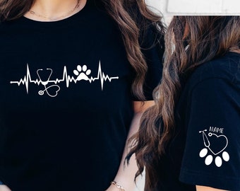 May include: Black t-shirt with a white graphic of a stethoscope, heart, and paw print inside a heartbeat line. The shirt also has a smaller graphic of a stethoscope, heart, and paw print on the sleeve. The text "NAME" is printed on the sleeve.