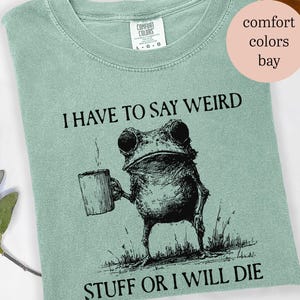 May include: A light green t-shirt with the text "I HAVE TO SAY WEIRD STUFF OR I WILL DIE" and a black and white illustration of a frog holding a mug. The shirt is a Comfort Colors brand.