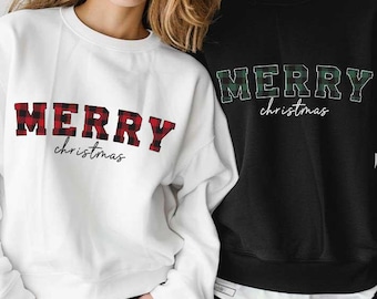 Puede incluir: Dos sudaderas, una blanca y otra negra, cada una con la inscripción "MERRY christmas" en la parte delantera. La sudadera blanca tiene letras a cuadros rojos y negros, mientras que la sudadera negra tiene letras a cuadros verdes y negros.