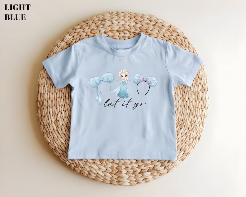 K&ouml;nnte beinhalten: Hellblaues T-Shirt mit einer Cartoon-Prinzessin-Grafik, zwei Micky-Maus-Ohren-Stirnb&auml;ndern und dem Schriftzug "let it go" in schwarzer Schrift. Das T-Shirt wird auf einer gewebten, kreisf&ouml;rmigen Matte pr&auml;sentiert.