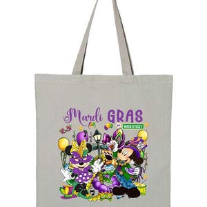 K&ouml;nnte beinhalten: Graue Tragetasche mit einem farbenfrohen Mardi Gras-Design mit Zeichentrickfiguren, Luftballons und dem Text "Mardi Gras on Main Street". Die Tasche hat einen hellgrauen Griff.