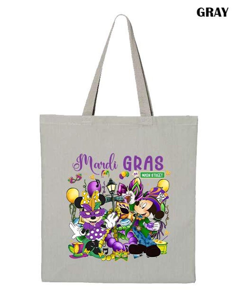 K&ouml;nnte beinhalten: Graue Tragetasche mit einem farbenfrohen Mardi Gras-Design mit Zeichentrickfiguren, Luftballons und dem Text "Mardi Gras on Main Street". Die Tasche hat einen hellgrauen Griff.