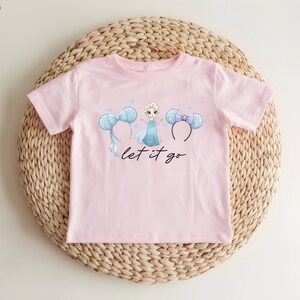 K&ouml;nnte beinhalten: Hellrosa T-Shirt mit einer Grafik einer Prinzessin, blauen Schleifen und dem Schriftzug "let it go" in schwarzer Schrift. Das Shirt wird auf einer gewebten, kreisf&ouml;rmigen Matte pr&auml;sentiert.