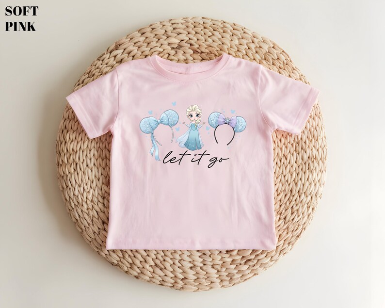 K&ouml;nnte beinhalten: Hellrosa T-Shirt mit einer Grafik einer Prinzessin, blauen Schleifen und dem Schriftzug "let it go" in schwarzer Schrift. Das Shirt wird auf einer gewebten, kreisf&ouml;rmigen Matte pr&auml;sentiert.