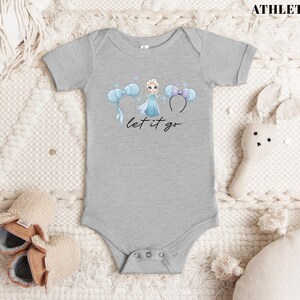 K&ouml;nnte beinhalten: Ein grauer Baby-Body mit einer Cartoon-Illustration einer Prinzessin in einem blauen Kleid, mit blauen und lila Micky-Maus-Ohren und dem Text "let it go". In der oberen rechten Ecke steht der Text "ATHLETIC H."