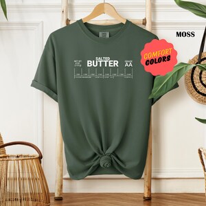 May include: A moss green t-shirt with the text "SALTED BUTTER" and a chart with the text "NET WT 1/2 LB (113g)" and "GRADE AA". The shirt has a knotted design in the front.