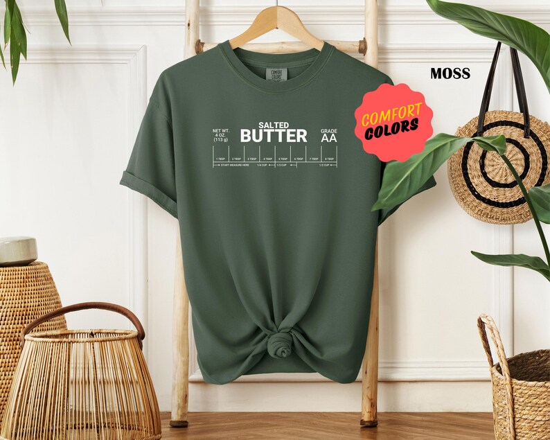 May include: A moss green t-shirt with the text "SALTED BUTTER" and a chart with the text "NET WT 1/2 LB (113g)" and "GRADE AA". The shirt has a knotted design in the front.