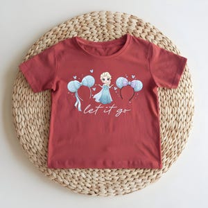 K&ouml;nnte beinhalten: Ein rotes T-Shirt mit einer Cartoon-Prinzessin in einem blauen Kleid, zwei Micky-Maus-Ohren-Haarb&auml;ndern und dem Text "let it go". Das T-Shirt liegt auf einer gewebten Matte.