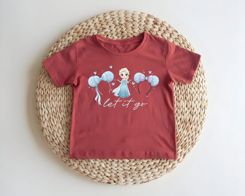 K&ouml;nnte beinhalten: Ein rotes T-Shirt mit einer Cartoon-Prinzessin in einem blauen Kleid, zwei Micky-Maus-Ohren-Haarb&auml;ndern und dem Text "let it go". Das T-Shirt liegt auf einer gewebten Matte.