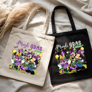 K&ouml;nnte beinhalten: Zwei Tragetaschen, eine beige und eine schwarze, mit Mardi-Gras-Designs. Die Taschen zeigen eine farbenfrohe Grafik von Zeichentrickfiguren in festlicher Kleidung mit den W&ouml;rtern "Mardi Gras" und "Main Street".