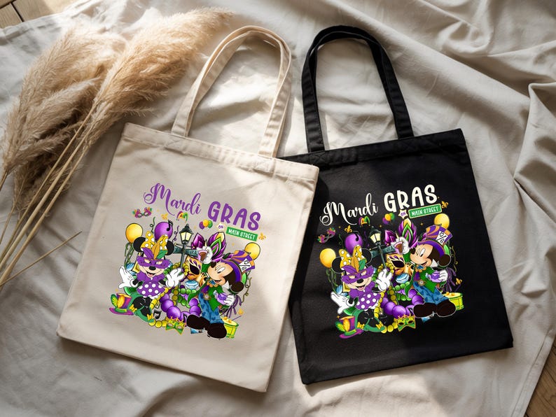 K&ouml;nnte beinhalten: Zwei Tragetaschen, eine beige und eine schwarze, mit Mardi-Gras-Designs. Die Taschen zeigen eine farbenfrohe Grafik von Zeichentrickfiguren in festlicher Kleidung mit den W&ouml;rtern "Mardi Gras" und "Main Street".