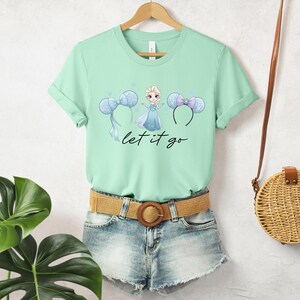 K&ouml;nnte beinhalten: Mintgr&uuml;nes T-Shirt mit einem Aufdruck einer Prinzessin und Micky-Maus-Ohren-Stirnb&auml;ndern. Der Text "let it go" steht darunter. Das T-Shirt wird mit Jeansshorts und einem geflochtenen G&uuml;rtel kombiniert. Rechts h&auml;ngt eine Strohtasche.
