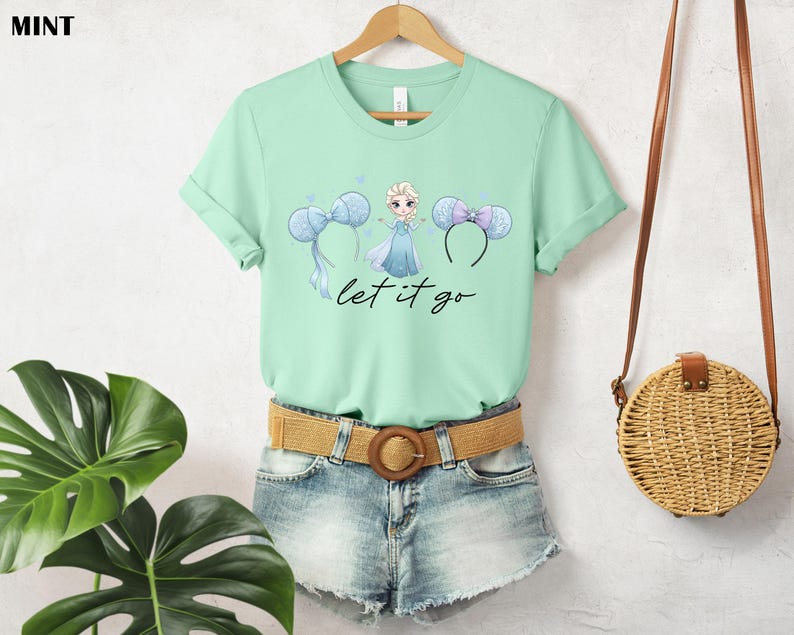 K&ouml;nnte beinhalten: Mintgr&uuml;nes T-Shirt mit einem Aufdruck einer Prinzessin und Micky-Maus-Ohren-Stirnb&auml;ndern. Der Text "let it go" steht darunter. Das T-Shirt wird mit Jeansshorts und einem geflochtenen G&uuml;rtel kombiniert. Rechts h&auml;ngt eine Strohtasche.