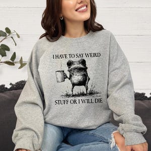 May include: A light gray sweatshirt with the text "I HAVE TO SAY WEIRD STUFF OR I WILL DIE." The graphic is a frog holding a mug. The sweatshirt is a casual, comfortable style.