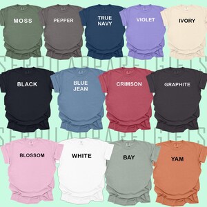 May include: A collection of 14 t-shirts in various colours, including moss green, pepper, true navy, violet, ivory, black, blue jean, crimson, graphite, blossom, white, bay, and yam. Each shirt has a different colour and a single word printed on the front.