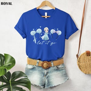 K&ouml;nnte beinhalten: K&ouml;nigsblaues T-Shirt mit einer Cartoon-Illustration einer Prinzessin in einem blauen Kleid und dem Text "let it go". Das Design umfasst blaue Ballons und Mauseohren-Stirnb&auml;nder. Das T-Shirt wird auf einem Holzb&uuml;gel pr&auml;sentiert.