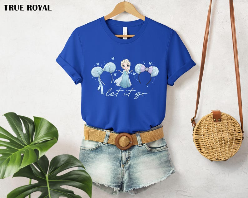K&ouml;nnte beinhalten: K&ouml;nigsblaues T-Shirt mit einer Cartoon-Illustration einer Prinzessin in einem blauen Kleid und dem Text "let it go". Das Design umfasst blaue Ballons und Mauseohren-Stirnb&auml;nder. Das T-Shirt wird auf einem Holzb&uuml;gel pr&auml;sentiert.