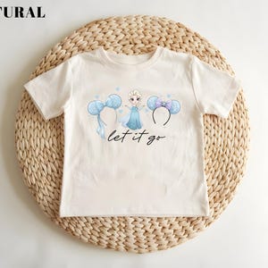 K&ouml;nnte beinhalten: Cremefarbenes T-Shirt mit einer Cartoon-Prinzessin-Grafik, Micky-Maus-Ohren-Haarb&auml;ndern und dem Schriftzug "let it go" in Schreibschrift. Das Shirt wird auf einer gewebten, kreisf&ouml;rmigen Matte pr&auml;sentiert.