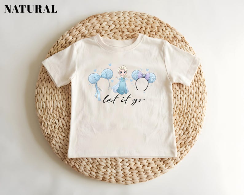 K&ouml;nnte beinhalten: Cremefarbenes T-Shirt mit einer Cartoon-Prinzessin-Grafik, Micky-Maus-Ohren-Haarb&auml;ndern und dem Schriftzug "let it go" in Schreibschrift. Das Shirt wird auf einer gewebten, kreisf&ouml;rmigen Matte pr&auml;sentiert.