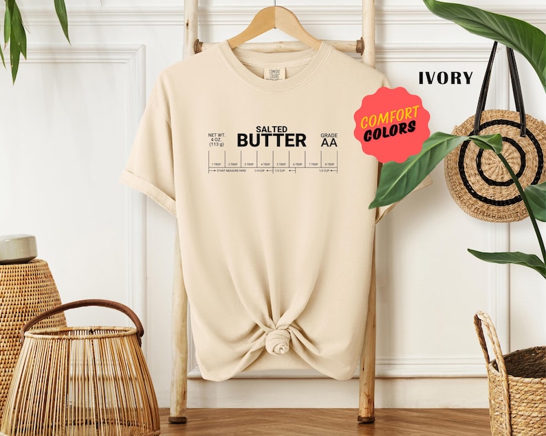 May include: A beige t-shirt featuring the text "SALTED BUTTER" and a graphic of a butter stick with measurements. The text "GRADE AA" is also on the shirt. The shirt is on a wooden hanger and has a knotted bottom.