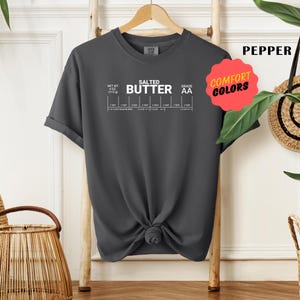 May include: A dark grey t-shirt with a white graphic of a salted butter label. The label includes the text "Salted Butter" and "Grade AA".