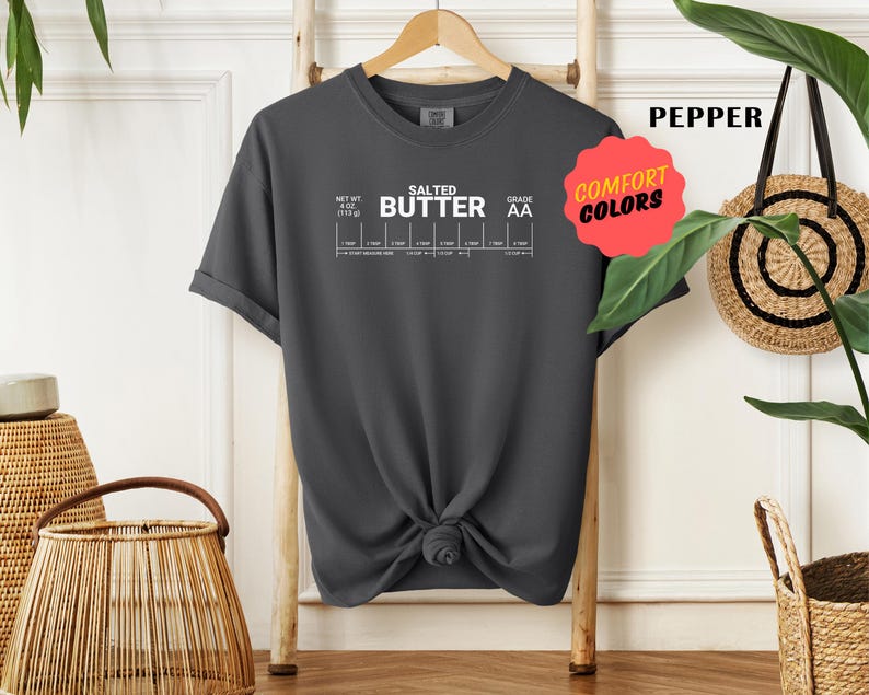 May include: A dark grey t-shirt with a white graphic of a salted butter label. The label includes the text "Salted Butter" and "Grade AA".