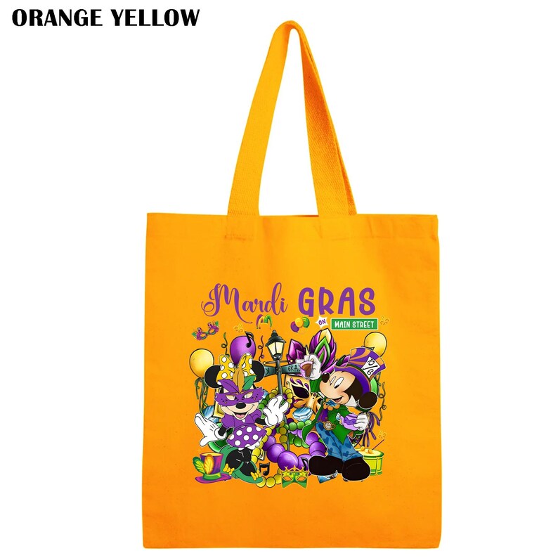K&ouml;nnte beinhalten: Orange-gelbe Tragetasche mit der Aufschrift "Mardi Gras on Main Street" und einer Grafik von Zeichentrickfiguren in festlicher Kleidung. Die Tasche hat lange Henkel und ist aus robustem Stoff gefertigt.