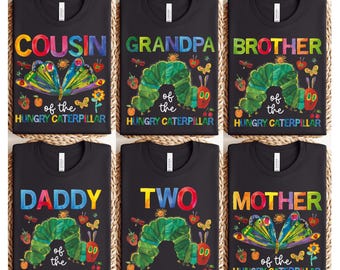 May include: Six black t-shirts with colourful graphics. Each shirt features text like "COUSIN," "GRANDPA," "BROTHER," "DADDY," "TWO," and "MOTHER." The designs include a green caterpillar, butterflies, and the text "of the HUNGRY CATERPILLAR."