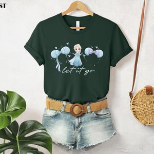 K&ouml;nnte beinhalten: Forstgr&uuml;nes T-Shirt mit einer Prinzessinnen-Illustration, blau-wei&szlig;en Luftballons und dem Schriftzug "let it go". Das T-Shirt wird mit Jeansshorts und einem geflochtenen G&uuml;rtel getragen. Das T-Shirt h&auml;ngt an einem Holzb&uuml;gel.