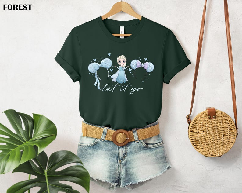 K&ouml;nnte beinhalten: Forstgr&uuml;nes T-Shirt mit einer Prinzessinnen-Illustration, blau-wei&szlig;en Luftballons und dem Schriftzug "let it go". Das T-Shirt wird mit Jeansshorts und einem geflochtenen G&uuml;rtel getragen. Das T-Shirt h&auml;ngt an einem Holzb&uuml;gel.