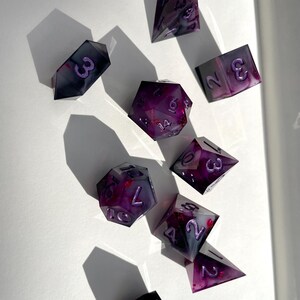 Könnte beinhalten: Ein Satz von zehn Würfeln in verschiedenen Formen, ideal für Tabletop-Spiele. Die Würfel sind eine Mischung aus Dunkelgrau und durchscheinendem Lila mit Marmoreffekt und lila Zahlen. Sie liegen auf einer weißen Oberfläche, mit Schatten.