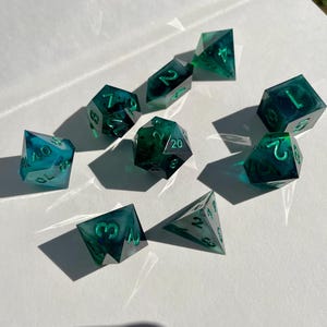 Könnte beinhalten: Ein Satz von zehn polyedrischen Würfeln in Türkis- und Smaragdgrüntönen. Die transparenten Würfel weisen interne Wirbel und Einschlüsse auf. Jeder Würfel hat weiße Zahlen. Die Würfel sind auf einer weißen Oberfläche angeordnet.