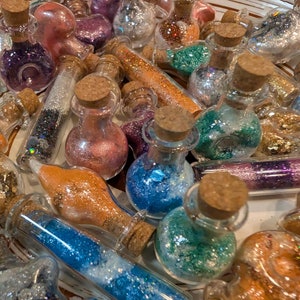 May include: A collection of small glass bottles and vials filled with colorful glitter. The bottles have cork stoppers and contain glitter in various colors, including blue, green, orange, purple, and silver. The items are arranged in a white bowl.