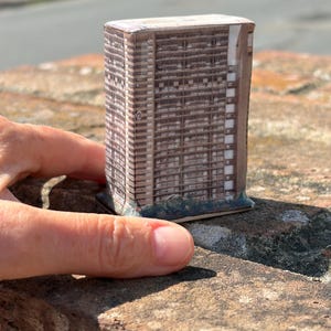 May include: A small, rectangular ceramic sculpture featuring a detailed photographic print of a tall, multi-storey brutalist apartment building with a grid of balconies and windows, rendered in shades of brown, grey, and white on a flat base.