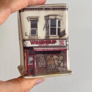 May include: A rectangular ceramic ornament featuring a photographic print of a storefront named WINEMEUP. The building facade includes two upper-story windows, a red storefront entrance, and a metal security shutter covered in graffiti art.