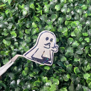 May include: A white glitter ghost sticker with a black outline and a peace sign hand gesture. The sticker is on a green background.
