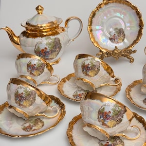 May include: A vintage tea set comprising a teapot, cups, and saucers. The set has a pearlescent finish with gold trim and handles. Each piece is decorated with a pastoral scene. Includes a decorative plate on a gold stand.