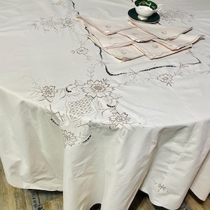 May include: A white tablecloth featuring intricate floral embroidery and cutwork details, accompanied by a stack of matching embroidered napkins and a single white and green teacup with a saucer placed on top of the folded fabric set.