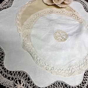 May include: A selection of antique white and cream doilies, featuring delicate lace and crochet work. The doilies are in various sizes and shapes, with scalloped edges and floral designs. A fabric flower is placed on top of the doilies.