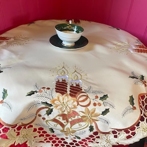 May include: A round, cream-coloured tablecloth featuring embroidered Christmas motifs including two red candles, golden bells, poinsettias, and holly leaves, with a scalloped, laser-cut lace border. A white and dark green teacup and saucer sit centred.