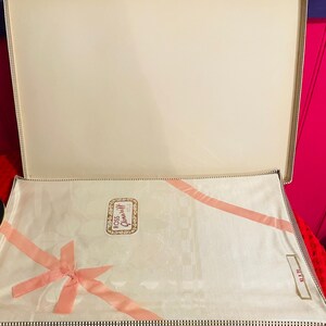May include: An open, cream-colored box with a white, floral-patterned cloth inside. A pink ribbon is tied in a bow and runs across the cloth. The box has a black and white checkered border.
