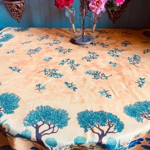 May include: A round tablecloth with a light orange background and a repeating pattern of teal trees and branches. The border features a wavy blue design. The tablecloth is draped over a table, with a vase of pink roses in the background.