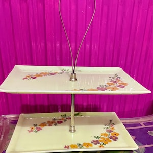 May include: Two-tiered serving tray with a silver metal handle and support. The rectangular trays are pale yellow and decorated with floral patterns in orange, purple, and yellow. The tray is designed for serving desserts or appetizers.