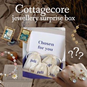 May include: A Cottagcore jewelry surprise box with the text "Chosen for you". The box contains small drawstring bags with the word "juli" printed on them. The image also shows a bracelet, earrings, and framed art.