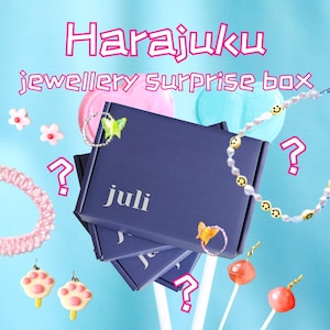 Könnte beinhalten: Eine marineblaue Schachtel mit dem Wort "juli" darauf, umgeben von Schmuck und Süßigkeiten. Die Schachtel ist mit "Harajuku jewellery SURPRISE box" in rosa Schrift beschriftet. Das Zubehör umfasst ein Armband, Ohrringe und eine Halskette.